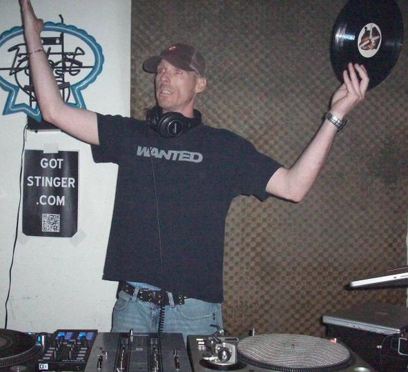 DJ Natural Nate® raising a vinyl record in the air at the decks